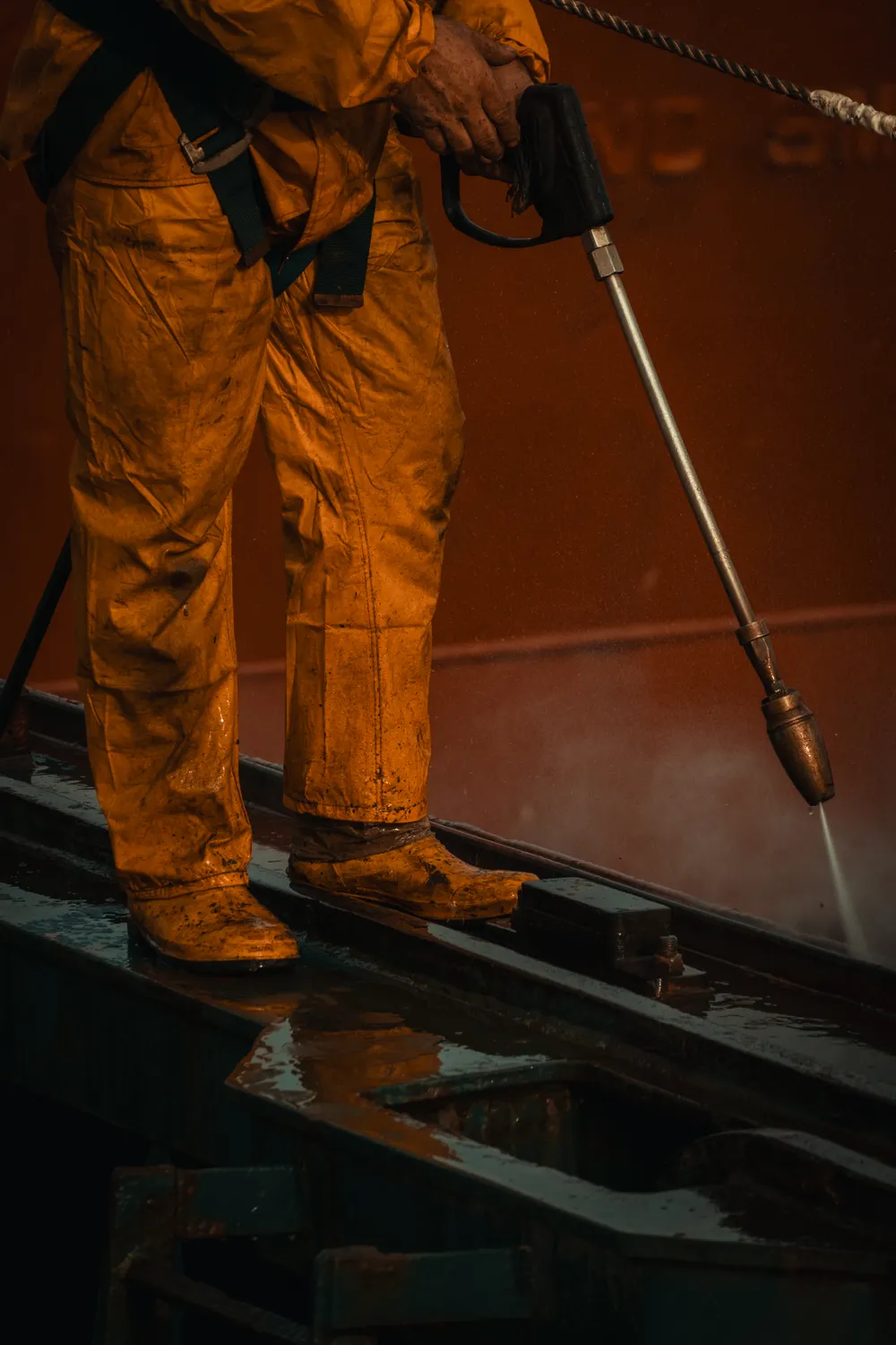 Pressure washer wand in gloved hands, water cutting across a steel surface